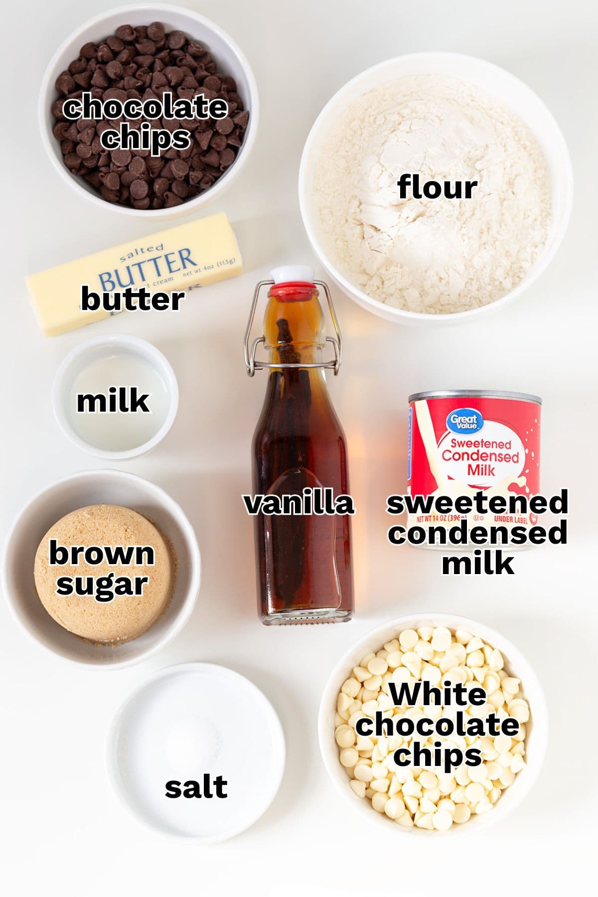 Top-down view of labeled baking ingredients in bowls and containers, perfect for making Cookie Dough Fudge: chocolate chips, flour, butter, milk, vanilla, sweetened condensed milk, brown sugar, white chocolate chips, and salt.