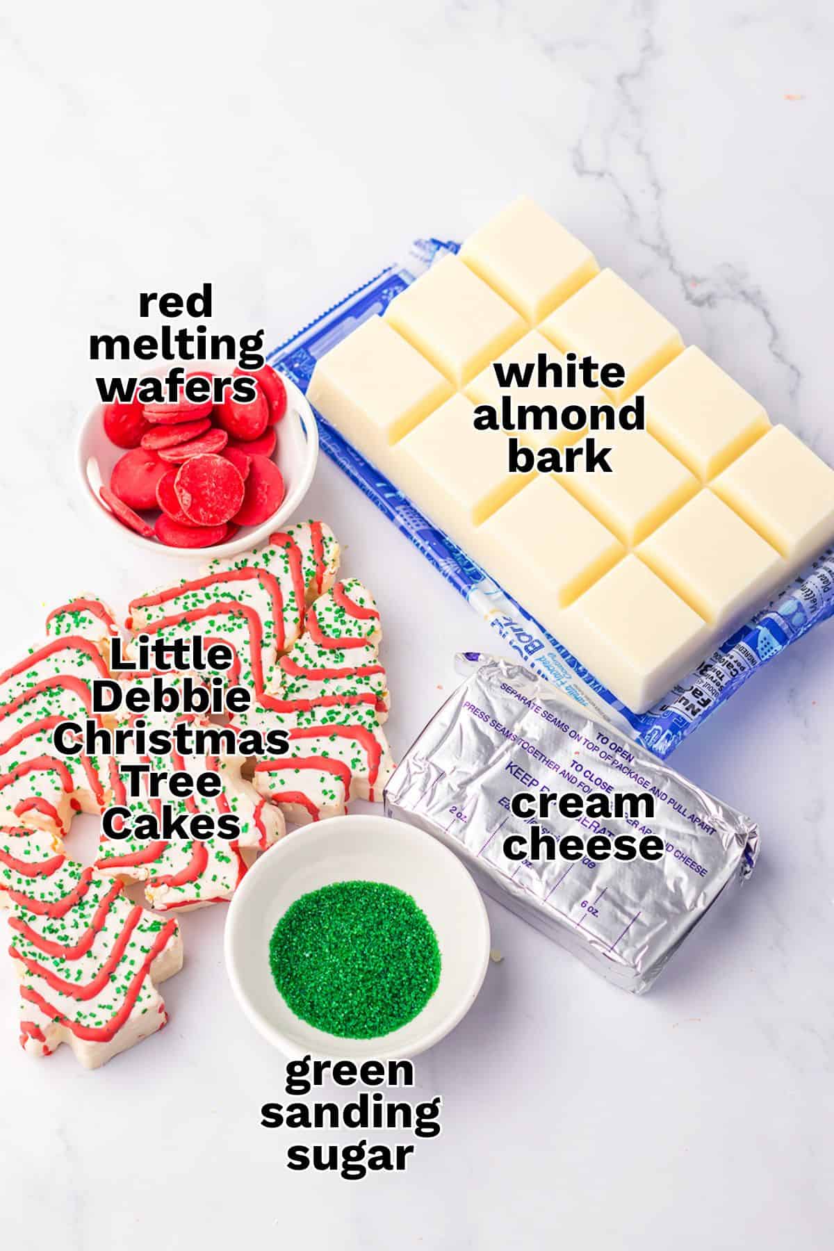 Ingredients for Christmas Tree Cake Balls are arranged on a counter: red melting wafers, white almond bark, Little Debbie Christmas tree cakes, cream cheese, and green sanding sugar.