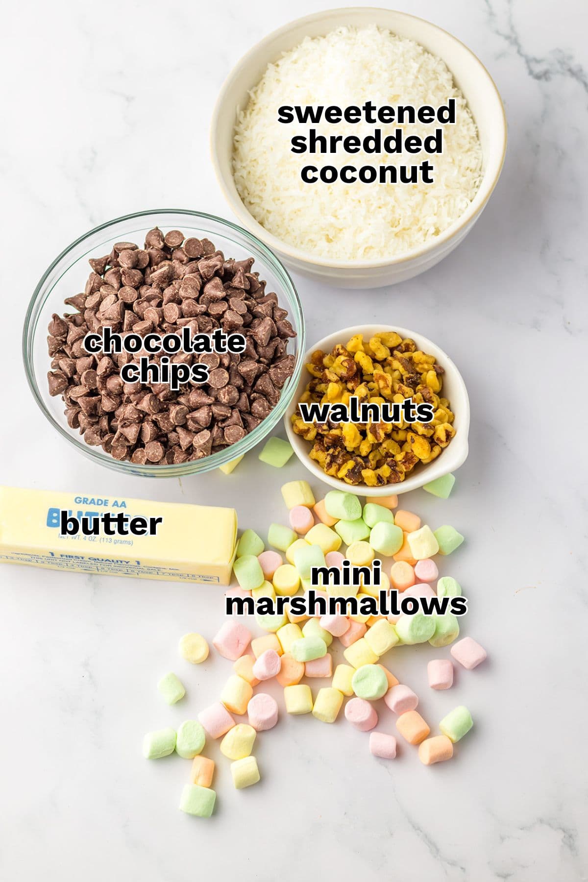 Ingredients for Church Window Cookies displayed on a white surface: sweetened shredded coconut, chocolate chips, walnuts, a stick of butter, and mini marshmallows—perfect for these easy no bake cookies.