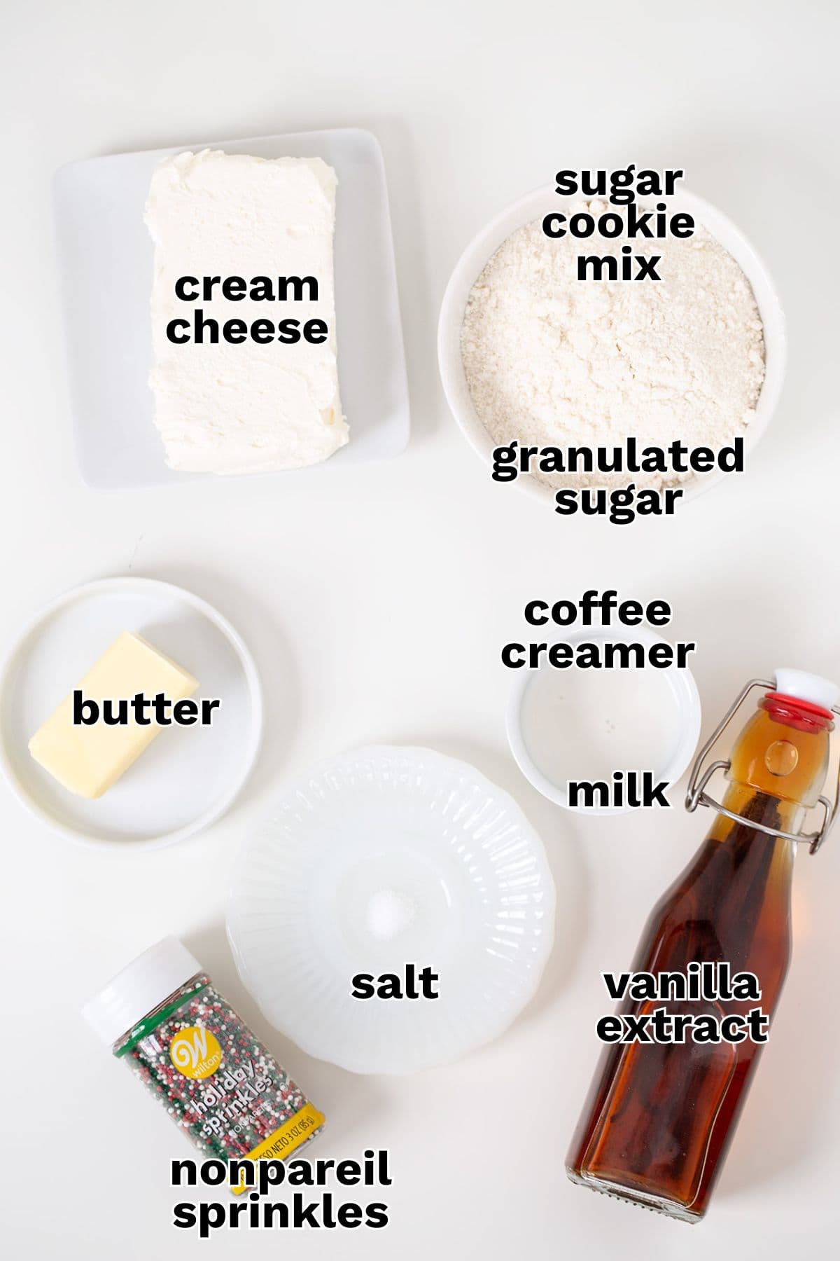 Various baking ingredients are arranged on a white surface, each labeled: cream cheese, sugar cookie mix, granulated sugar, coffee creamer, butter, milk, salt, vanilla extract, and nonpareil sprinkles—perfect for making a delicious Sugar Cookie Dip.