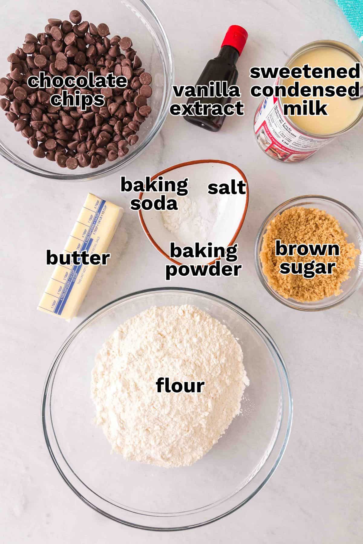 A countertop displays chocolate chips, vanilla extract, sweetened condensed milk, butter sticks, and other essentials in separate bowls&mdash;everything you need to bake irresistible chocolate chip cookies.