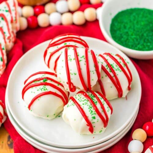 Five white chocolate-coated Christmas Tree Cake Balls with red drizzle and green sprinkles are arranged on a white plate; green sprinkles and festive decor are in the background.