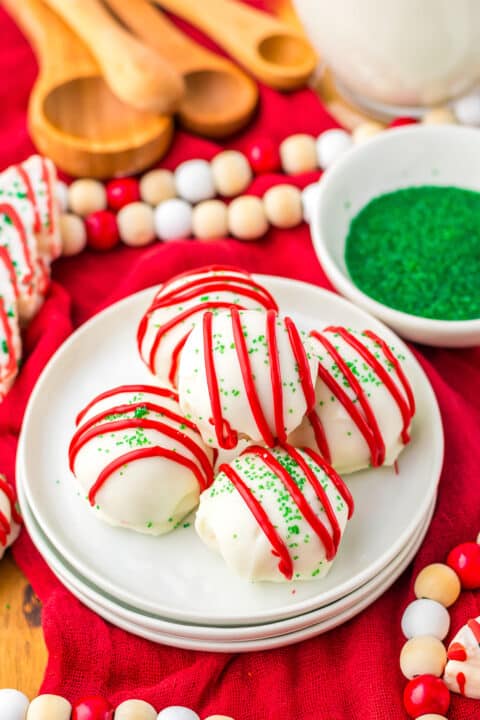 Four Christmas Tree Cake Balls, inspired by Little Debbie, are coated in white chocolate with red icing and green sprinkles on a white plate, surrounded by festive decorations and a bowl of green sprinkles.