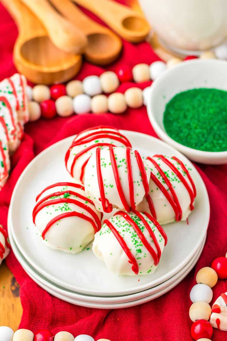 Four Christmas Tree Cake Balls, inspired by Little Debbie, are coated in white chocolate with red icing and green sprinkles on a white plate, surrounded by festive decorations and a bowl of green sprinkles.