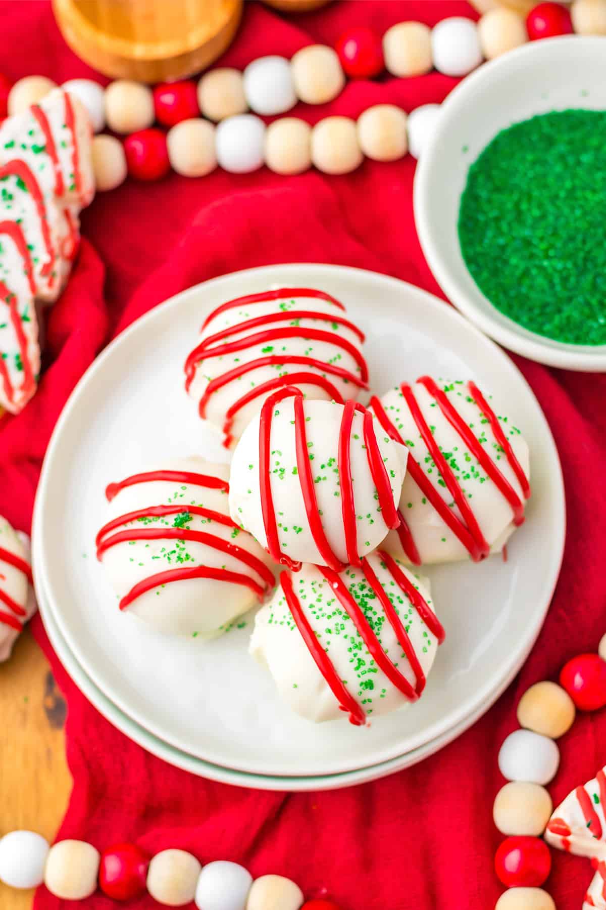 White chocolate-covered Christmas Tree Cake Balls with red icing drizzle and green sprinkles are arranged on a white plate, surrounded by festive decorations and a bowl of green sprinkles.