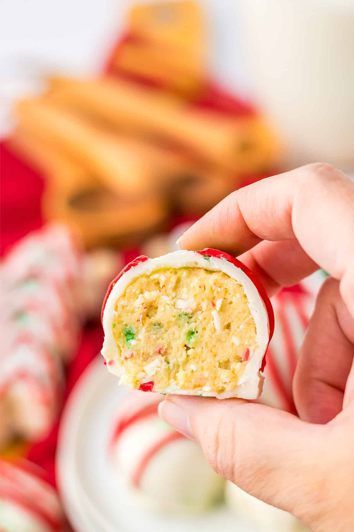 A hand holds a white chocolate-coated Cake Ball with a yellow interior and colorful sprinkles, with more Little Debbie treats and truffles blurred in the background.