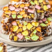 Three slices of Church Window Cookies with colorful mini marshmallows and coconut are stacked on a plate, with more no bake cookies in the background.