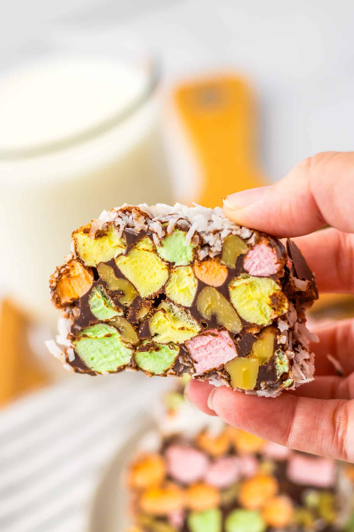 A hand holds a slice of No Bake Church Window Cookies, made with colorful marshmallows, chocolate, nuts, and shredded coconut.