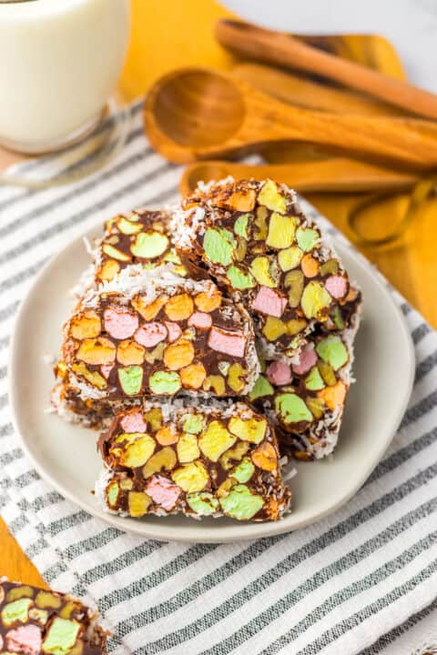 A plate of chocolate-covered Church Window Cookies with colorful mini marshmallows sits on a striped cloth, wooden measuring spoons in the background—perfect for fans of classic no bake cookies.