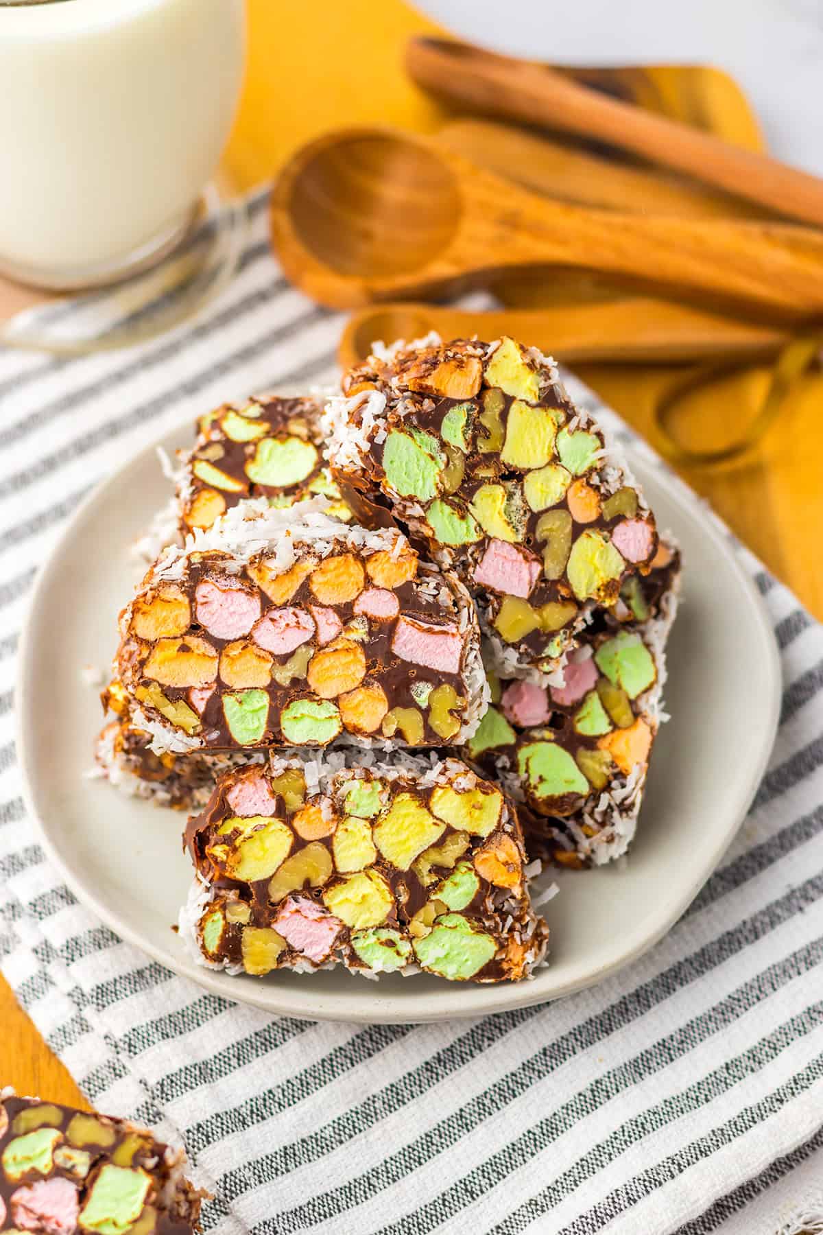 A plate of chocolate-covered Church Window Cookies with colorful mini marshmallows sits on a striped cloth, wooden measuring spoons in the background—perfect for fans of classic no bake cookies.
