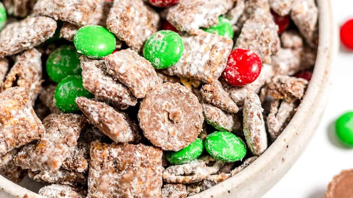 A bowl filled with chocolate-covered cereal pieces dusted in powdered sugar, mixed with red and green Christmas Candies for a festive touch.