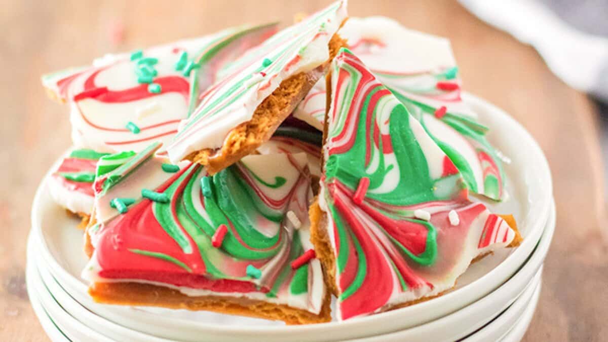 A stack of holiday-themed peppermint bark pieces with red, green, and white swirled chocolate and sprinkles sits on a white plate&mdash;perfect for adding to cookie boxes or sharing as homemade Christmas candies.