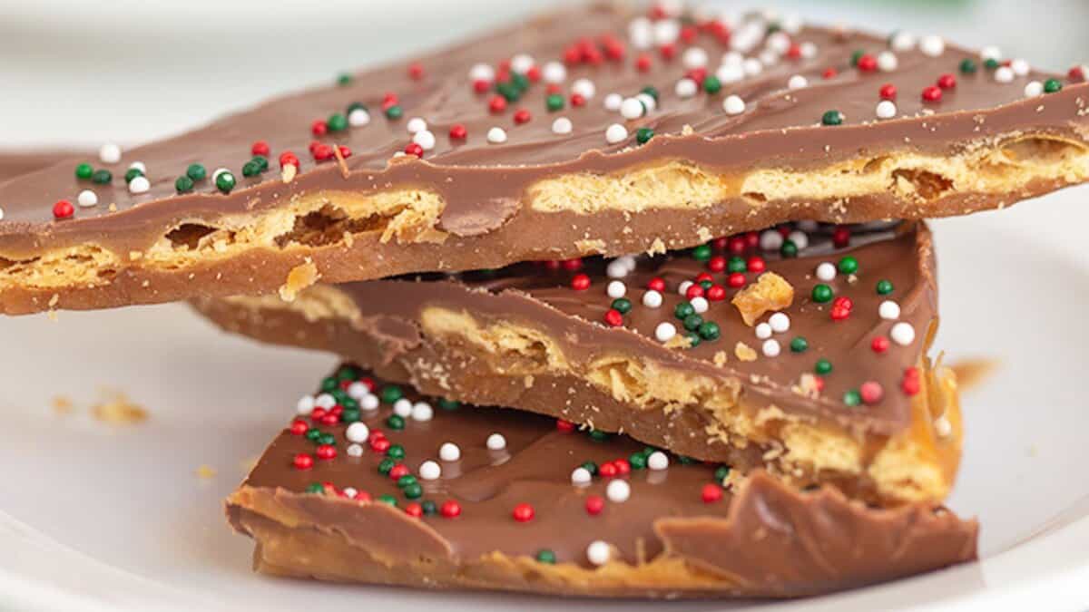 Three pieces of chocolate toffee bark with red, white, and green sprinkles are stacked on a white plate&mdash;perfect for adding to your cookie boxes or sharing as homemade Christmas candies.