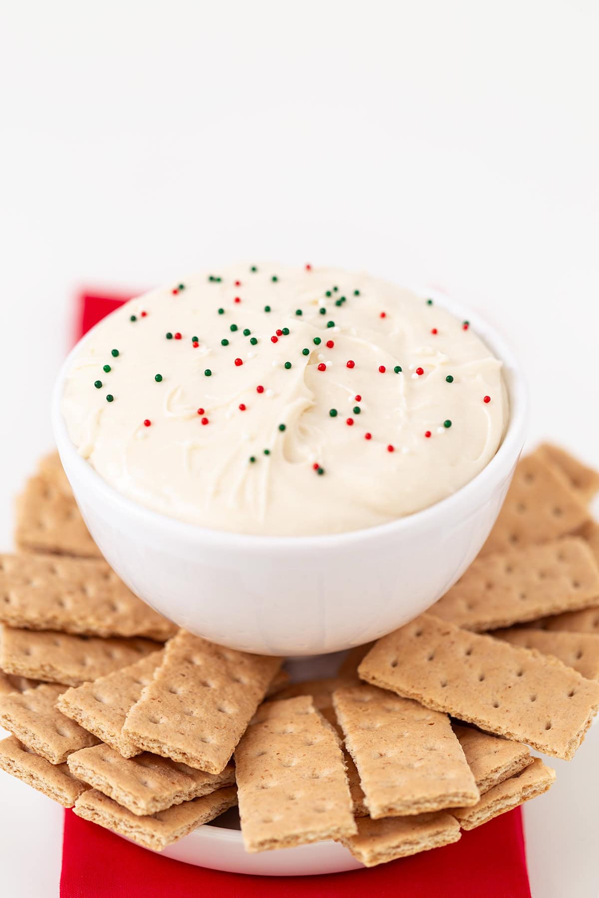 A white bowl filled with creamy Sugar Cookie Dip topped with red and green sprinkles, surrounded by rectangular graham crackers on a white plate.