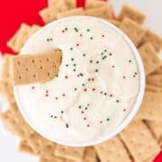 A bowl of creamy Sugar Cookie Dip topped with red and green sprinkles, surrounded by rectangular graham crackers on a red napkin.