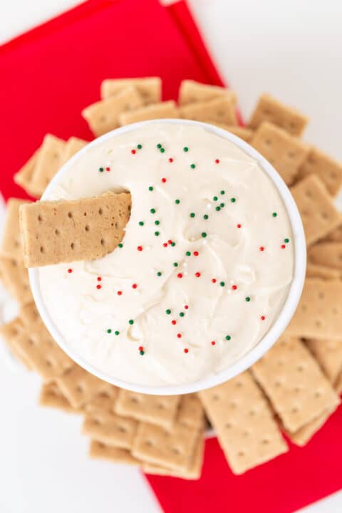 A bowl of creamy Sugar Cookie Dip topped with red and green sprinkles, surrounded by rectangular graham crackers on a red napkin.