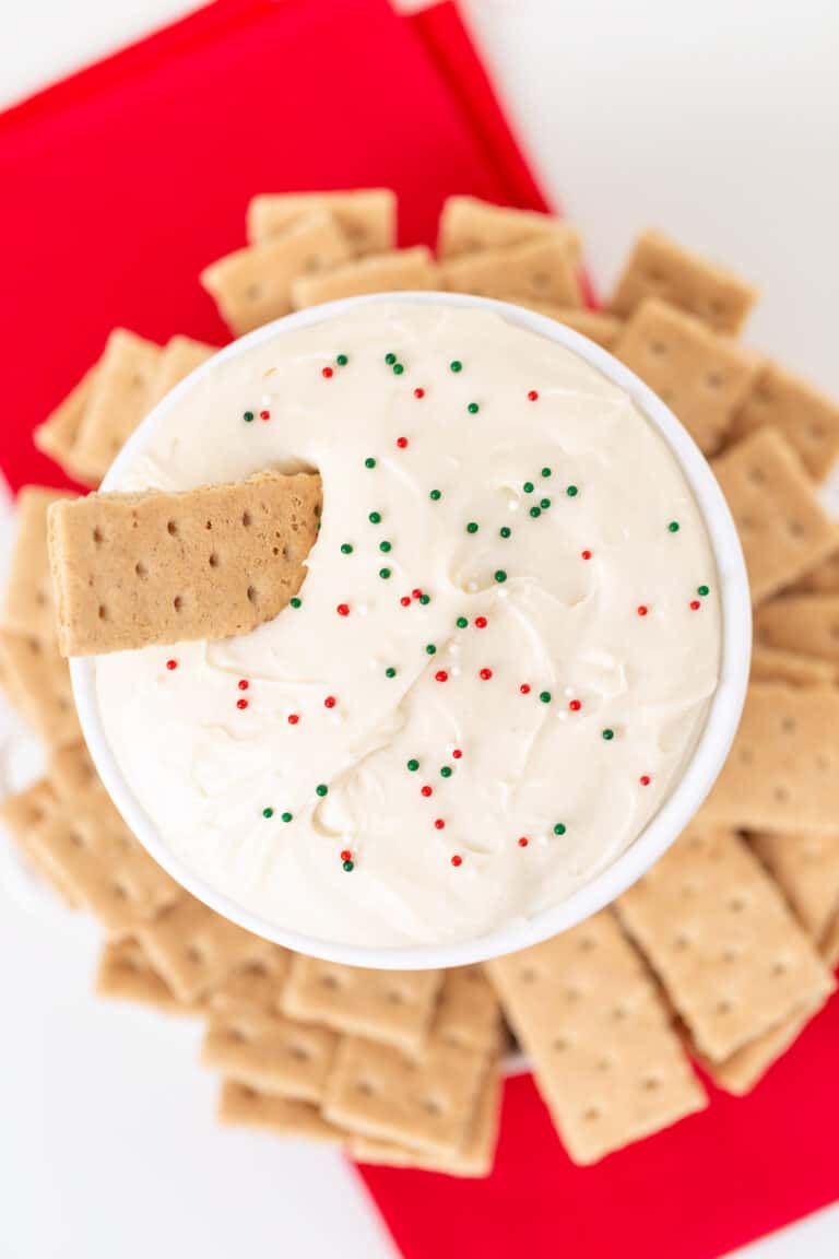 A bowl of creamy Sugar Cookie Dip topped with red and green sprinkles, surrounded by rectangular graham crackers on a red napkin.