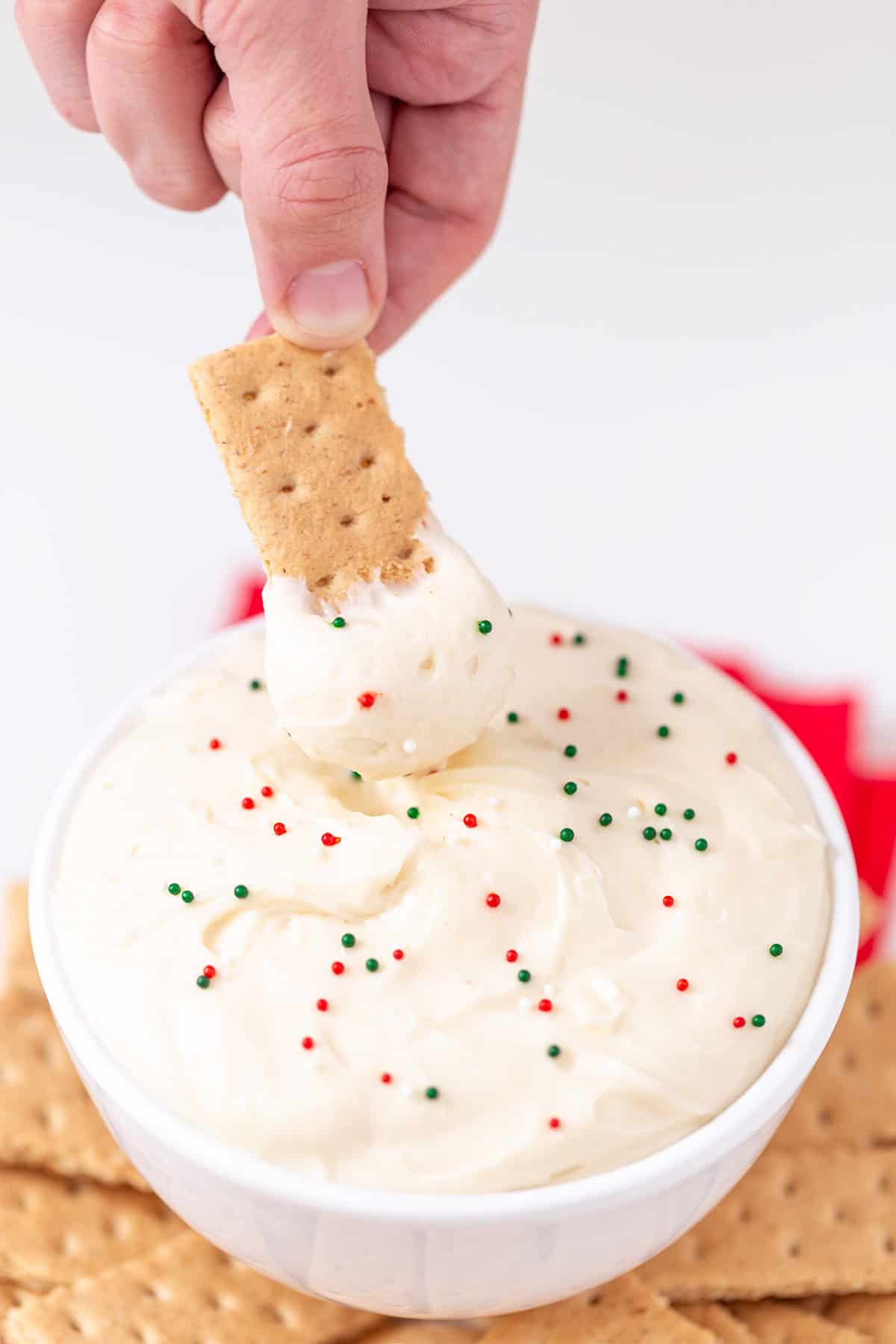 A hand dips a graham cracker into a bowl of creamy Sugar Cookie Dip, topped with festive red and green sprinkles.