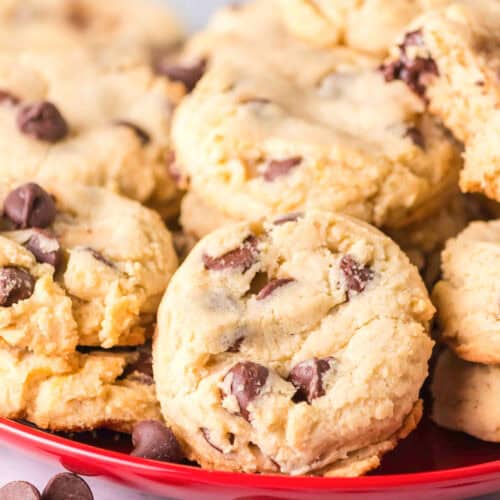 A red plate holds a pile of chocolate chip cookies made with sweetened condensed milk, with a few chocolate chips scattered on the white surface nearby.