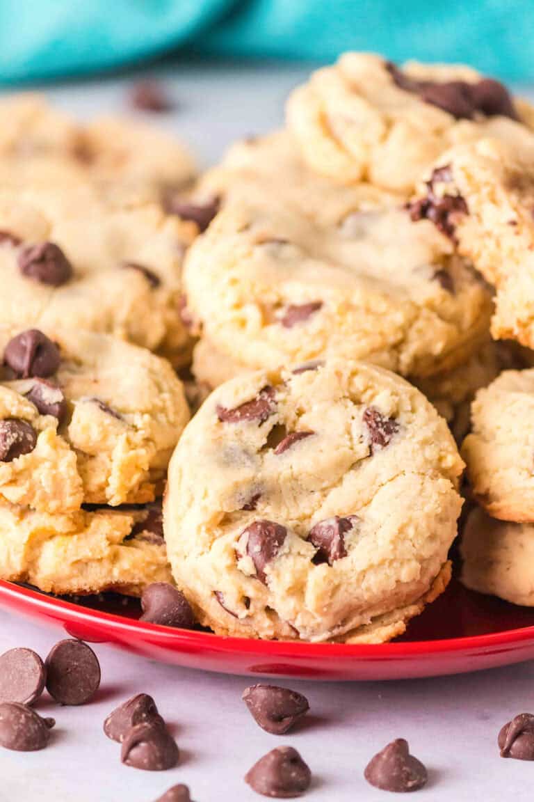 A red plate holds a pile of chocolate chip cookies made with sweetened condensed milk, with a few chocolate chips scattered on the white surface nearby.