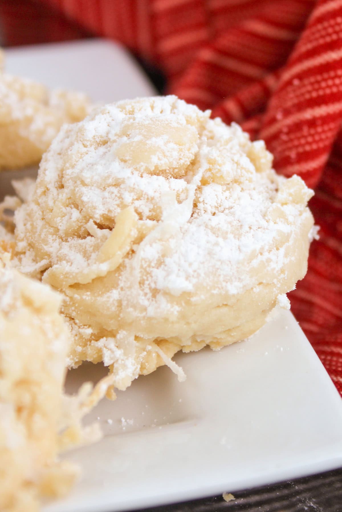 A round snowball cookie, made with sweetened condensed milk and dusted with powdered sugar, sits on a white plate near a red textured cloth.
