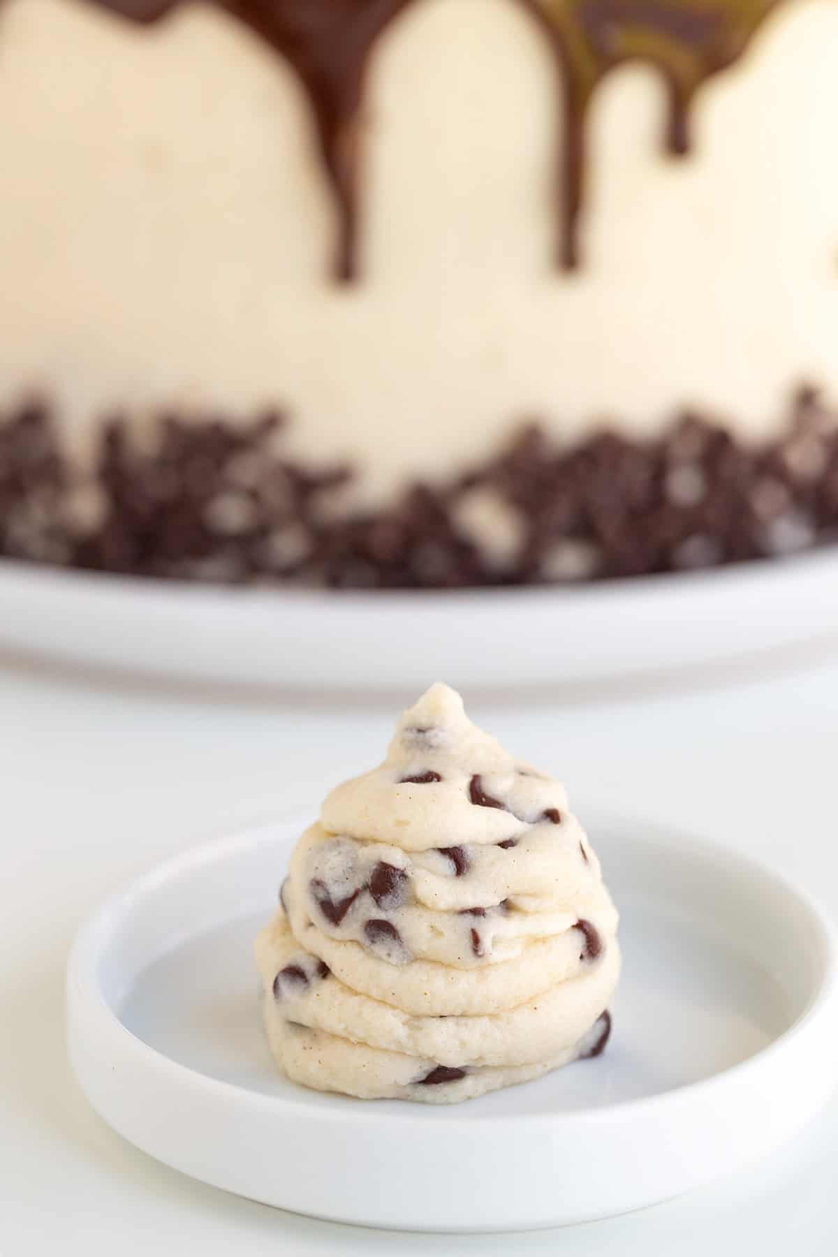 A single swirl of chocolate chip cookie dough sits on a small white plate, with a frosted cake decorated with chocolate, chocolate chips, and luscious Cannoli Filling in the background.