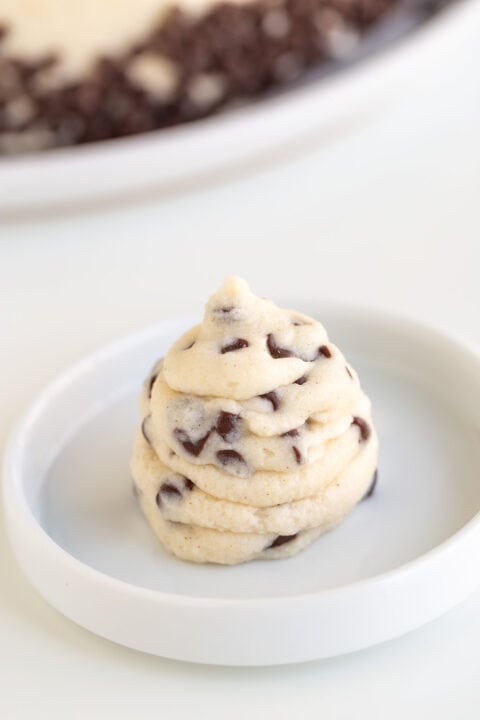 A swirl of raw chocolate chip cookie dough, infused with creamy Cannoli Filling, sits on a white plate, with a blurred plate of more dough in the background.