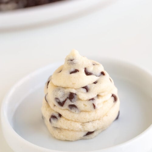 A swirl of raw chocolate chip cookie dough, infused with creamy Cannoli Filling, sits on a white plate, with a blurred plate of more dough in the background.