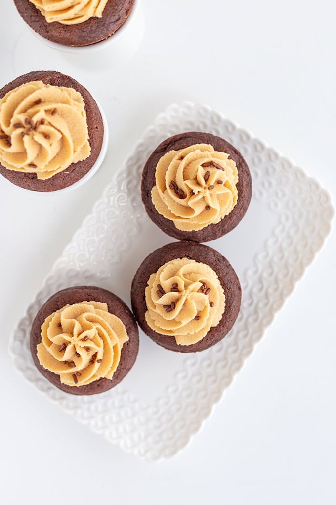 Four chocolate cupcakes topped with peanut butter cream cheese frosting and chocolate sprinkles are arranged on a white decorative tray against a white background.