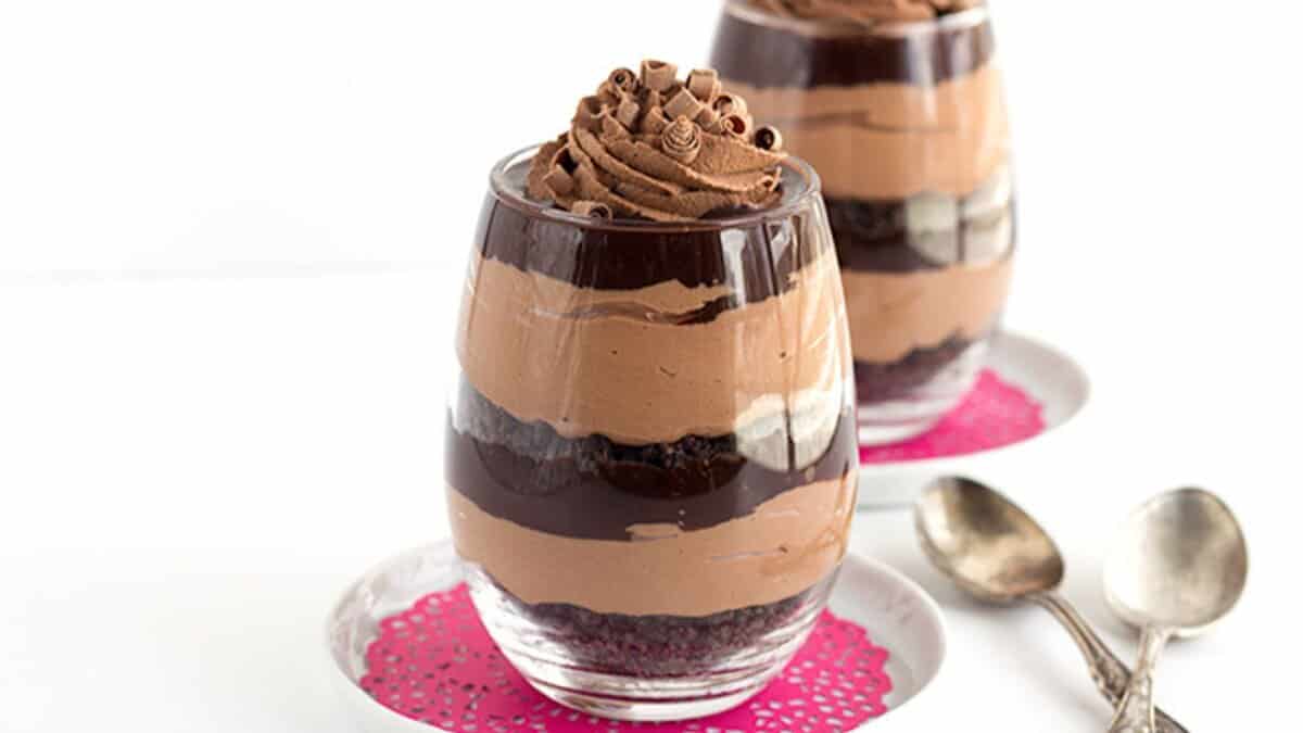 A glass cup filled with layered chocolate mousse, whipped cream, and chocolate shavings&mdash;one of the most decadent desserts&mdash;sits on a white plate with a pink doily, next to two metal spoons.