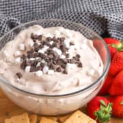 A glass bowl of Hot Cocoa Dip—a delicious dessert dip with chocolate chips and mini marshmallows—sits on a wooden board with graham crackers and fresh strawberries nearby.