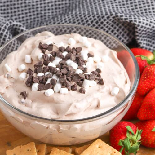 A glass bowl of Hot Cocoa Dip&mdash;a delicious dessert dip with chocolate chips and mini marshmallows&mdash;sits on a wooden board with graham crackers and fresh strawberries nearby.