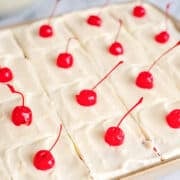 A Cherry Sheet Cake sits on a baking sheet, each square piece topped with smooth white frosting and a bright red maraschino cherry.