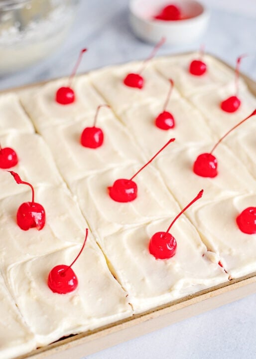 A Cherry Sheet Cake sits on a baking sheet, each square piece topped with smooth white frosting and a bright red maraschino cherry.