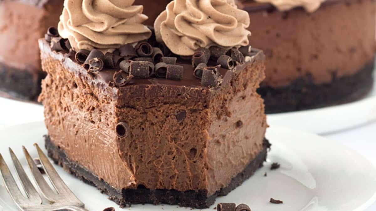 A slice of chocolate cheesecake with a chocolate cookie crust, topped with whipped cream and chocolate curls, sits on a white plate with a fork&mdash;perfect for fans of decadent desserts and irresistible chocolate recipes.