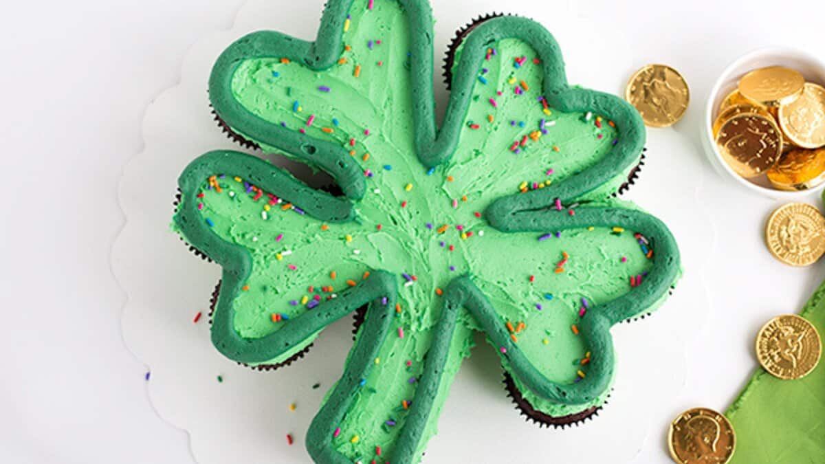 A clover-shaped cake with green frosting and colorful sprinkles sits next to gold chocolate coins in a bowl, making it the perfect St. Patrick's Day treat for fans of mint chocolate desserts.