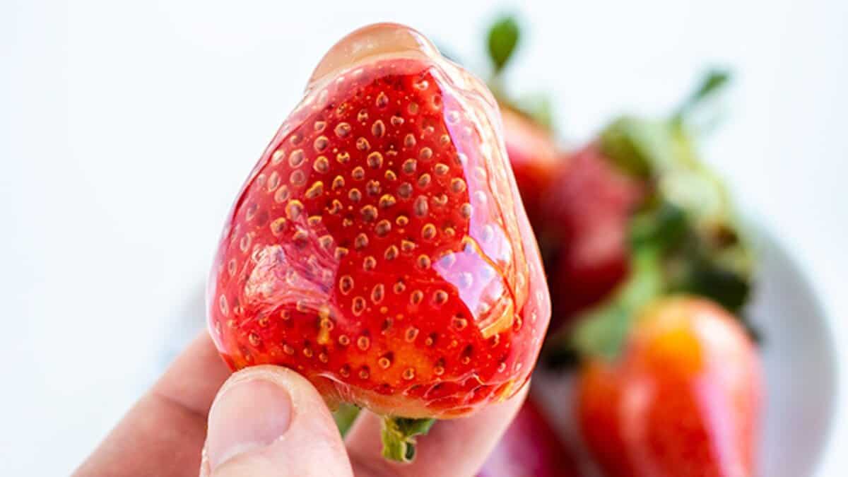 A hand holds a strawberry covered in a clear, gelatinous layer, with more fresh strawberries blurred in the background—capturing the essence of spring desserts.