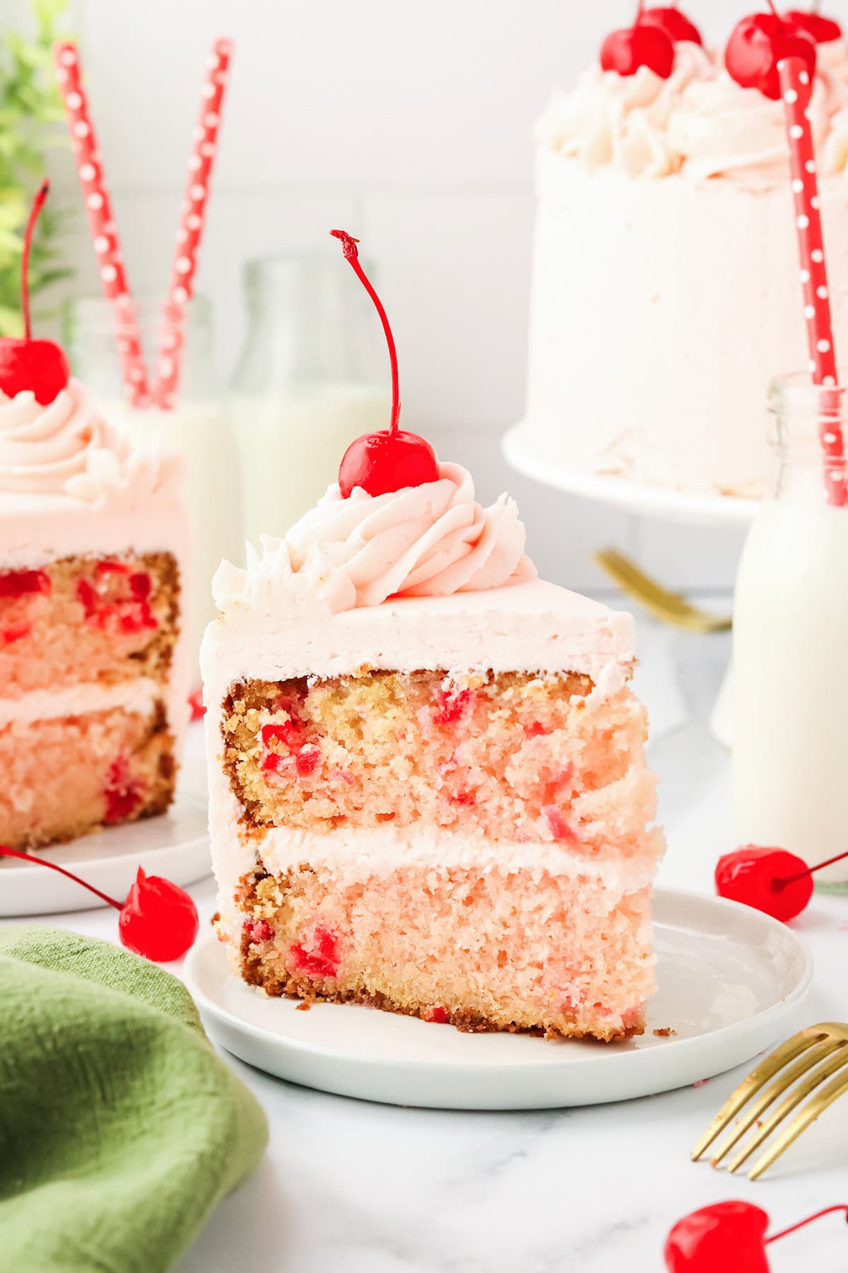 A slice of homemade cherry chip cake with pink frosting and a cherry on top, served on a white plate, with milk and more cake in the background.