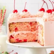 A sliced Cherry Chip Cake with pink frosting and maraschino cherries on top sits on a white cake stand.