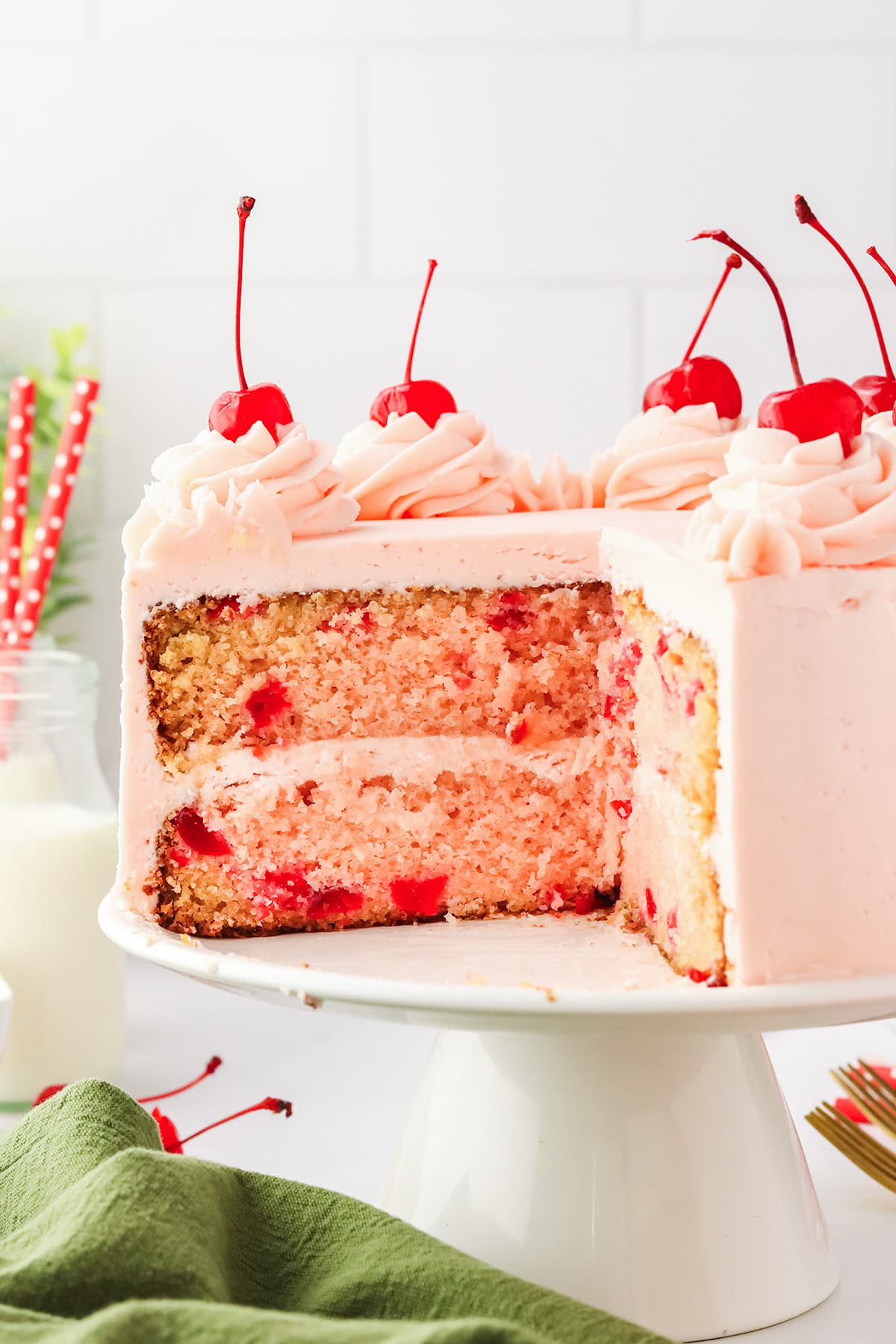 A sliced Cherry Chip Cake with pink frosting and maraschino cherries on top sits on a white cake stand.