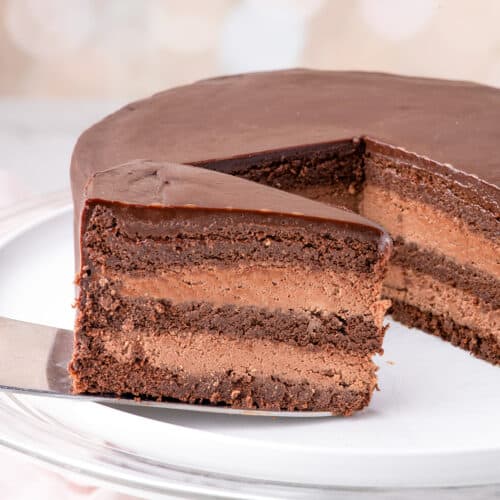 A luscious Chocolate Mousse Cake with layers of rich cake and glossy chocolate frosting, one slice being lifted from the whole cake on a glass cake stand.