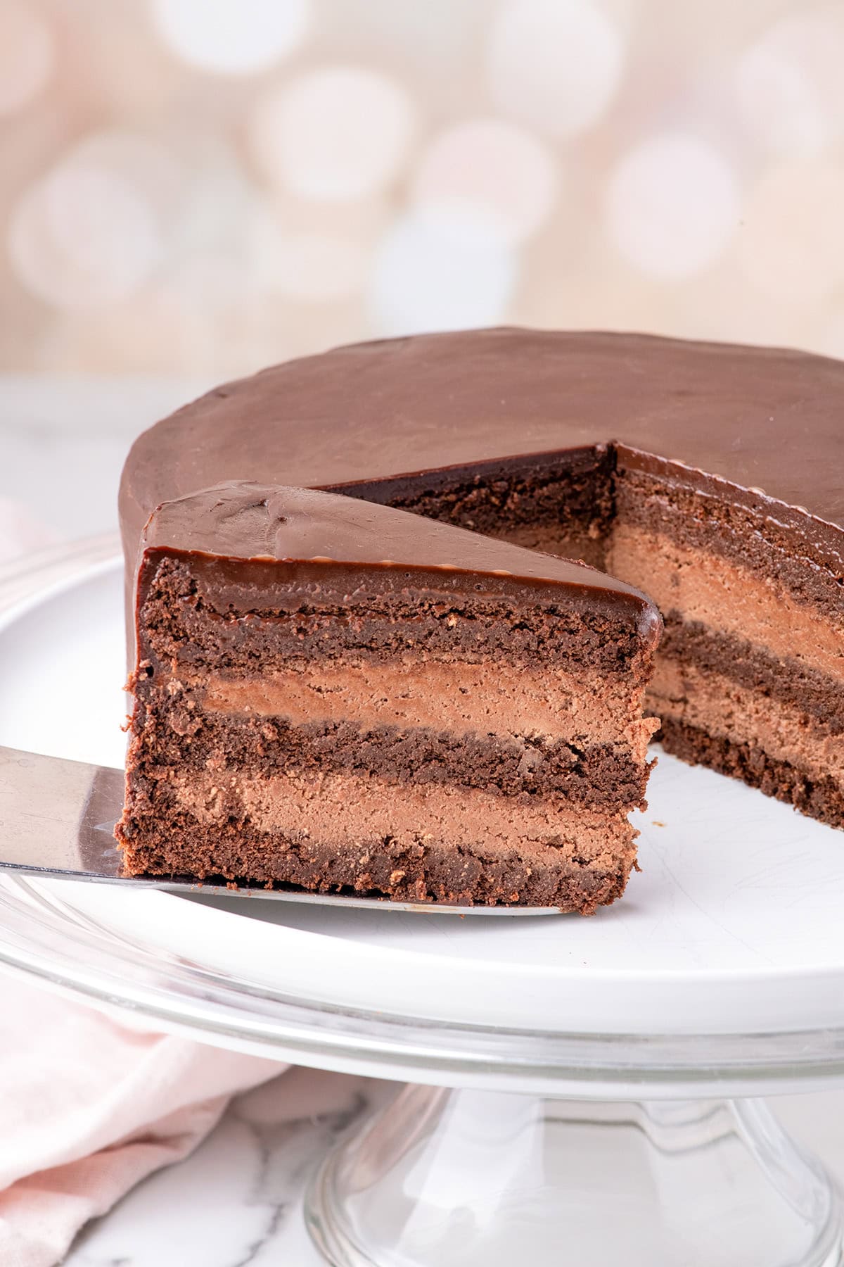 A luscious Chocolate Mousse Cake with layers of rich cake and glossy chocolate frosting, one slice being lifted from the whole cake on a glass cake stand.
