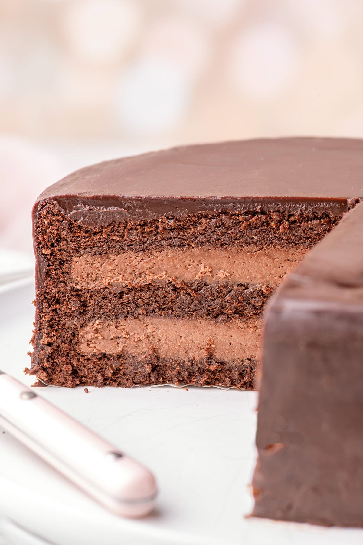 A sliced Chocolate Mousse Cake with rich chocolate frosting and filling, showing multiple decadent layers on a white plate with a knife beside it.