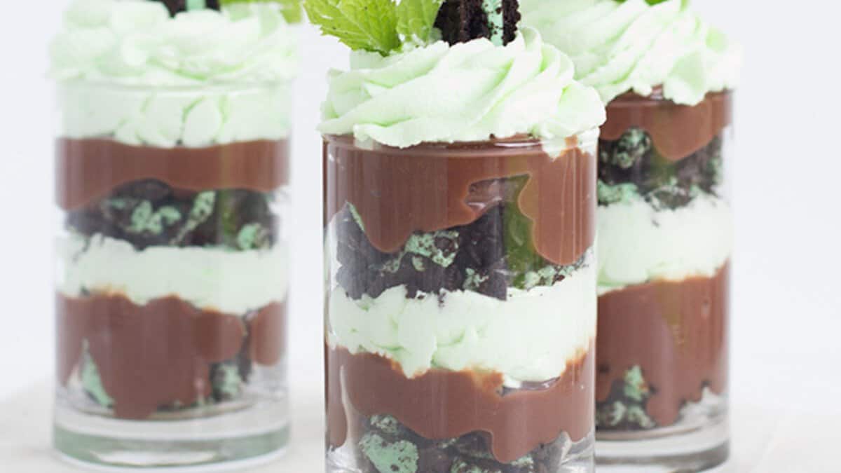 Three glass cups filled with layered chocolate pudding, mint cream, and crushed chocolate cookies create the perfect Mint Chocolate Desserts for St. Patrick's Day, topped with whipped cream and a fresh mint leaf.