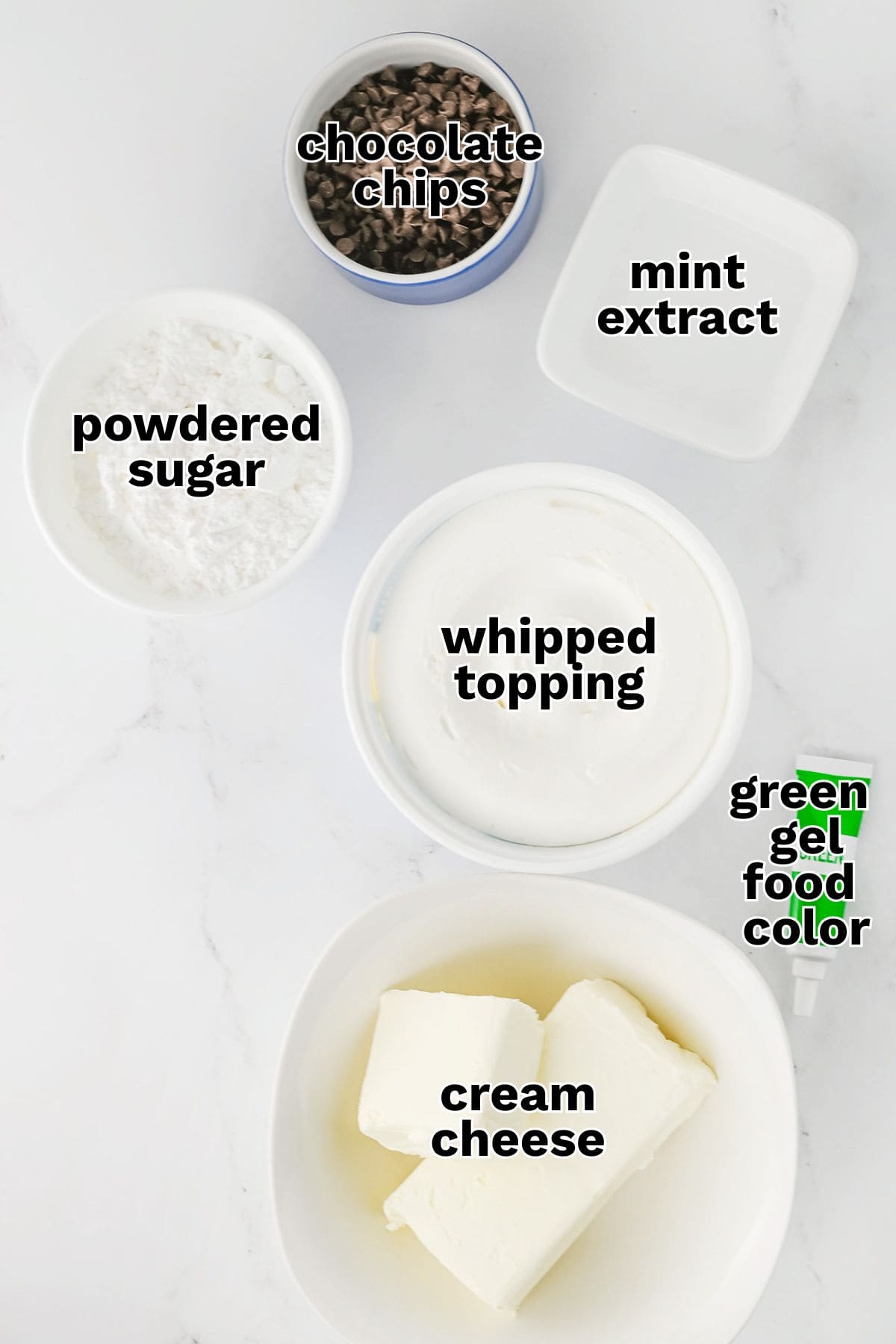 Five labeled ingredients for Mint Chocolate Chip Dip on a marble surface: chocolate chips, mint extract, powdered sugar, whipped topping, green gel food color, and cream cheese.