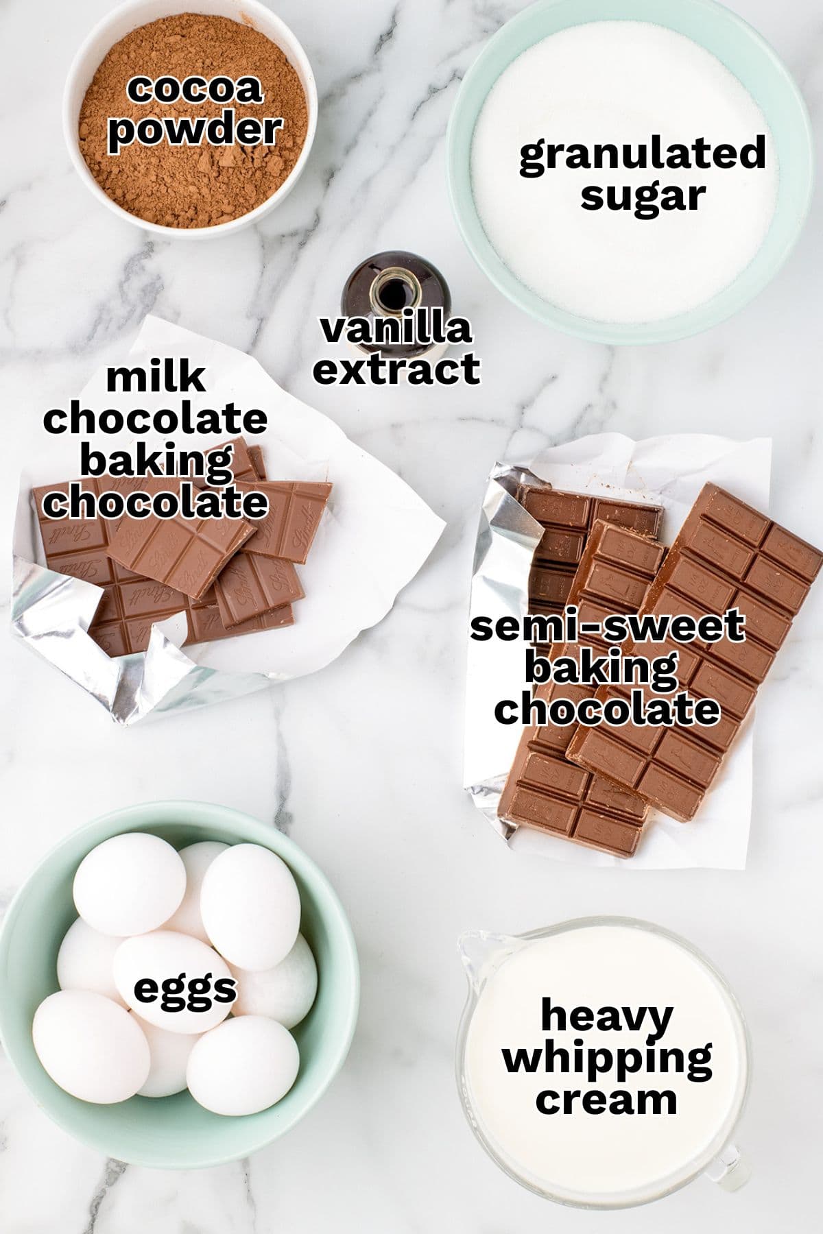 Top-down view of ingredients for Chocolate Mousse Cake on a marble surface, including cocoa powder, granulated sugar, vanilla extract, milk and semi-sweet baking chocolate, eggs, and heavy whipping cream.