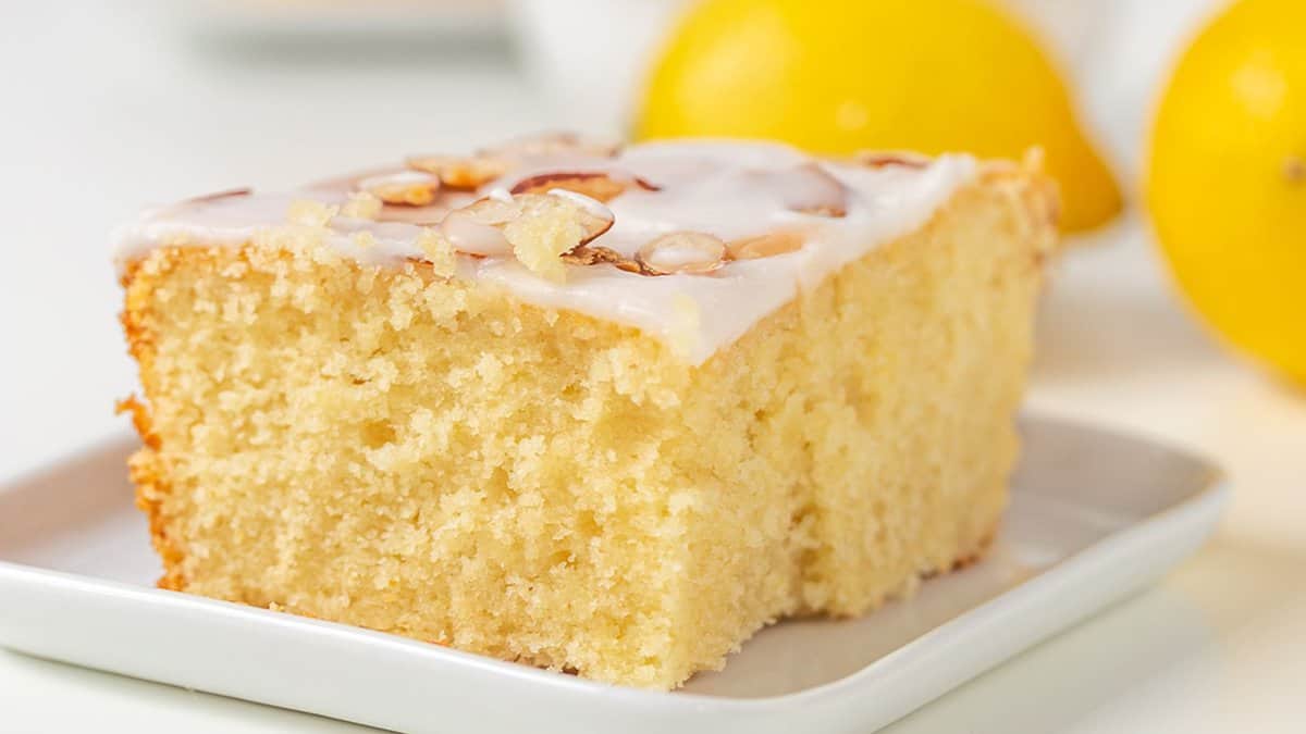 A square slice of yellow lemon cake with icing and almond slices on top, placed on a white plate with lemons in the background—perfect for anyone who loves bright, refreshing lemon desserts.