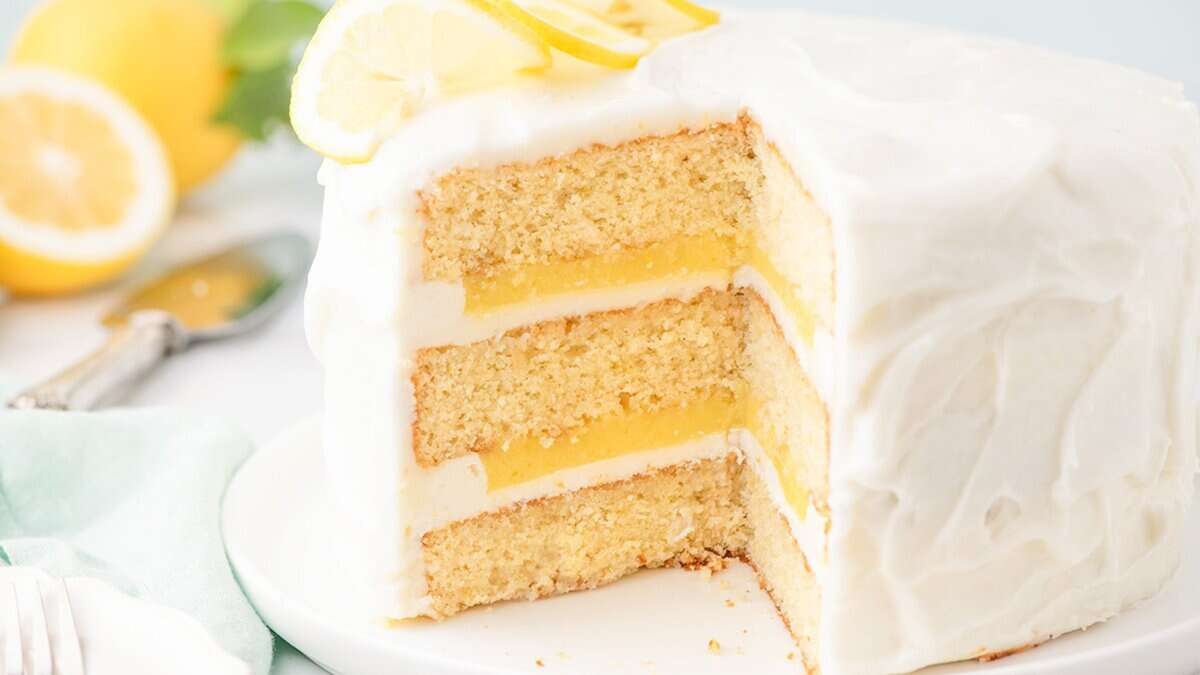 Three-layer lemon cake with white frosting and lemon curd filling, topped with lemon slices, on a white plate—perfect for fans of lemon desserts or anyone seeking fresh spring desserts. Sliced lemons in the background add a vibrant touch.