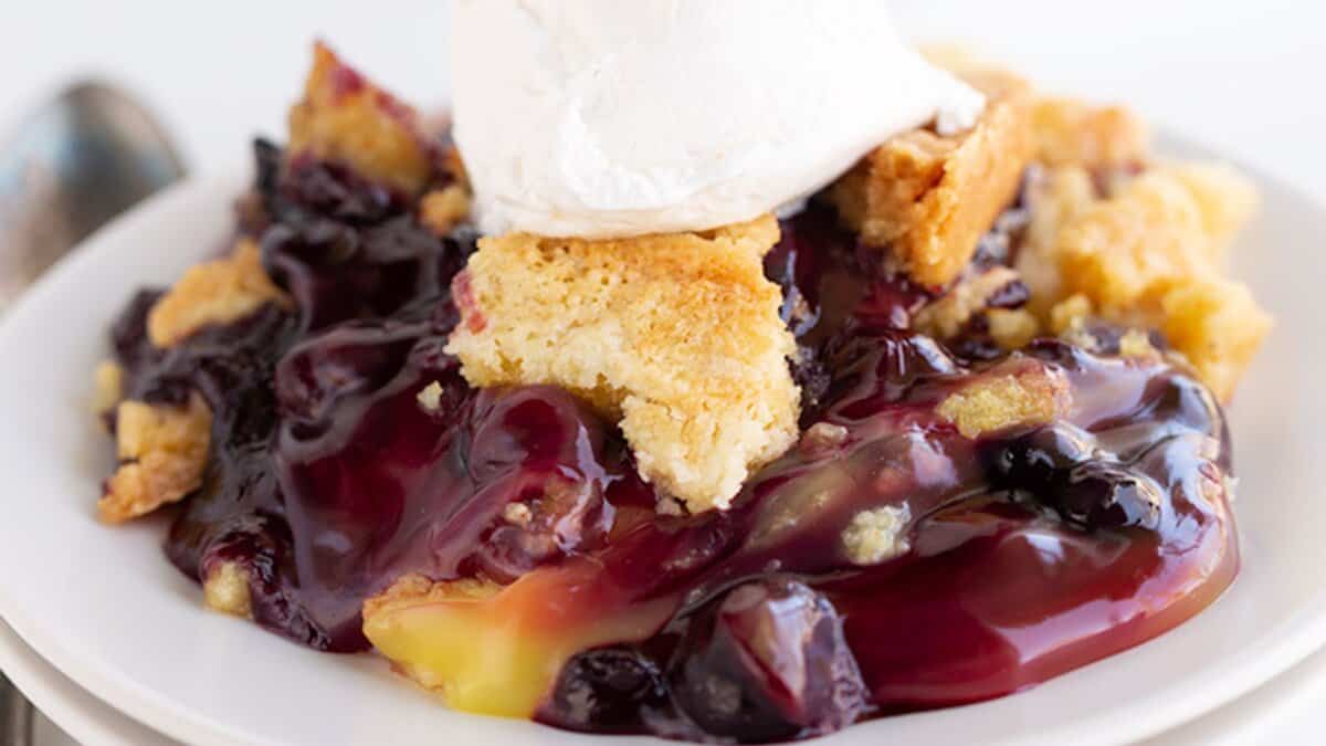 A serving of blueberry cobbler with a golden crust and a dollop of whipped cream on top, presented on a white plate—perfect for fans of spring desserts.