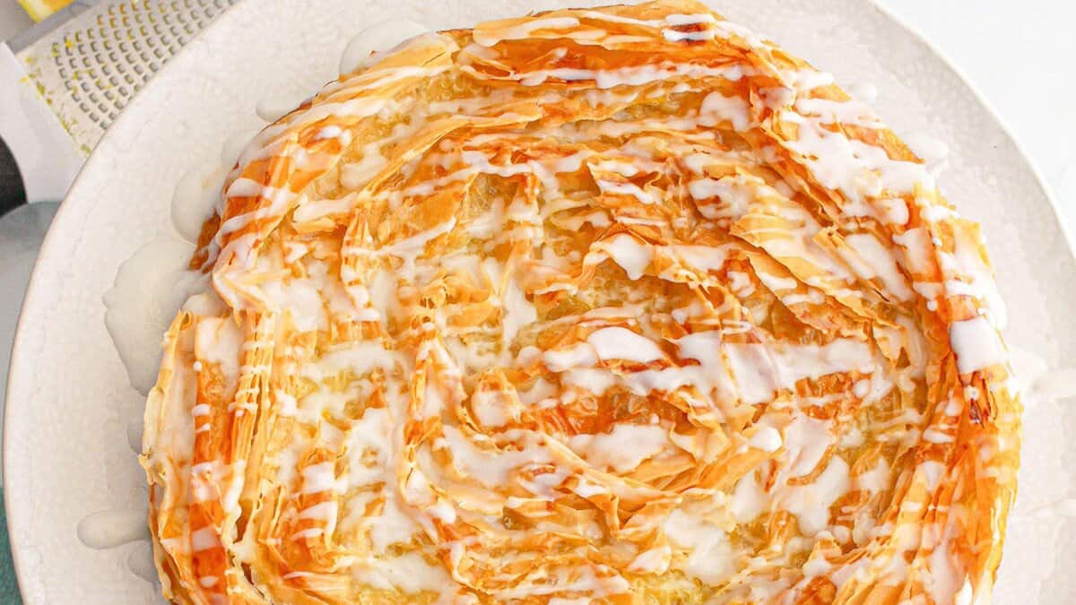 A round pastry with flaky, golden layers is drizzled with white icing and displayed on a white plate, making it a delightful choice for fans of lemon desserts and spring desserts.